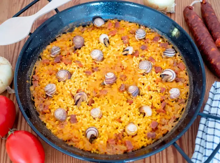 Arroz con caracoles en paellera, con trozos de chorizo y decorado con caracoles. Ingredientes frescos alrededor.